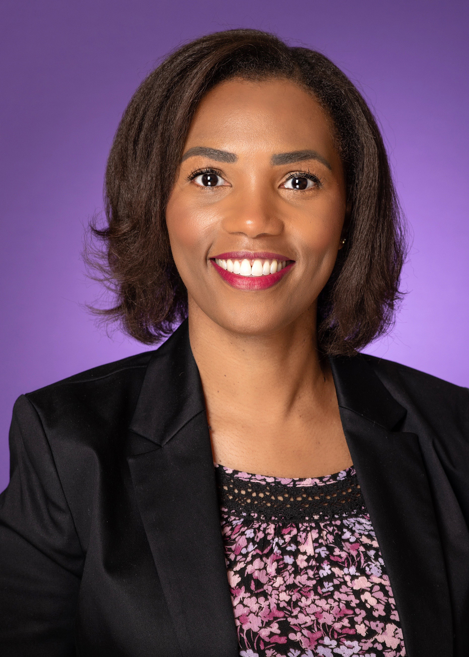 A woman smiles, wearing a multi-colored shirt, black blazer and a purple background.