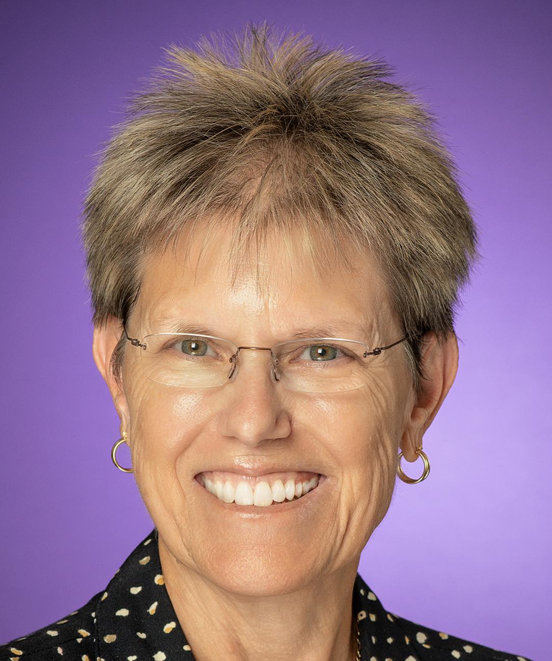 A woman with short hair, smiling, wearing glasses with a purple background.