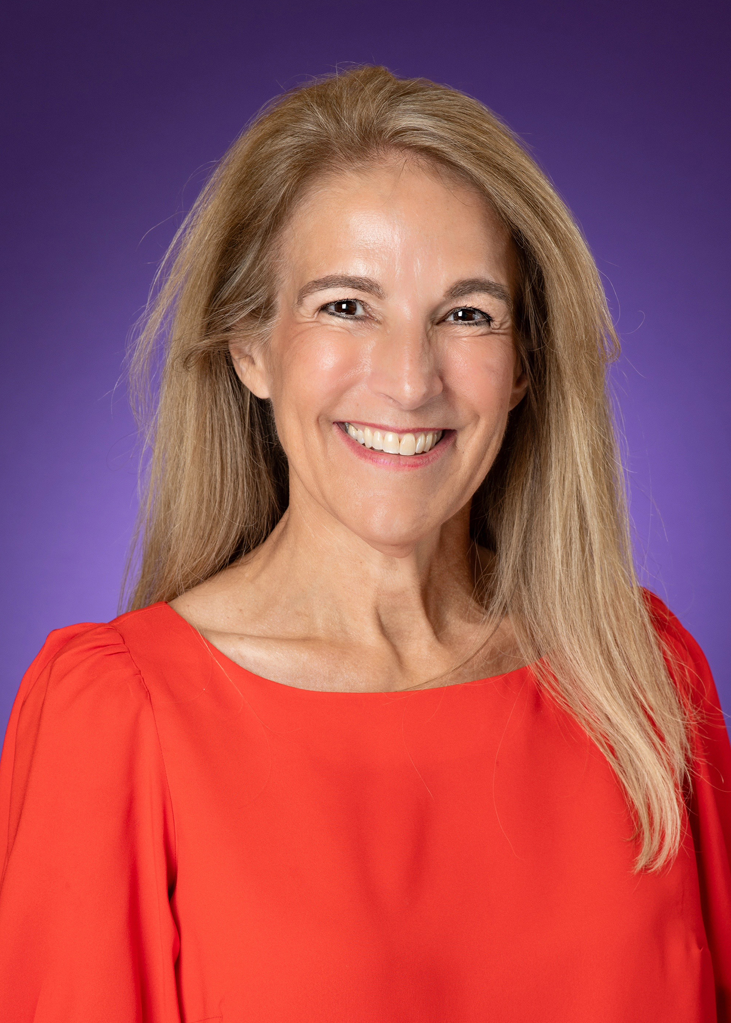 A woman smiles, wearing a red-orange shirt and a purple background.