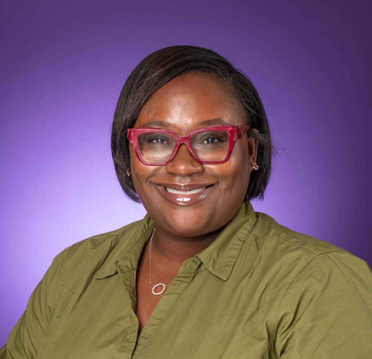 A photo of a woman smiling, wearing red glasses and a green shirt with a purple background