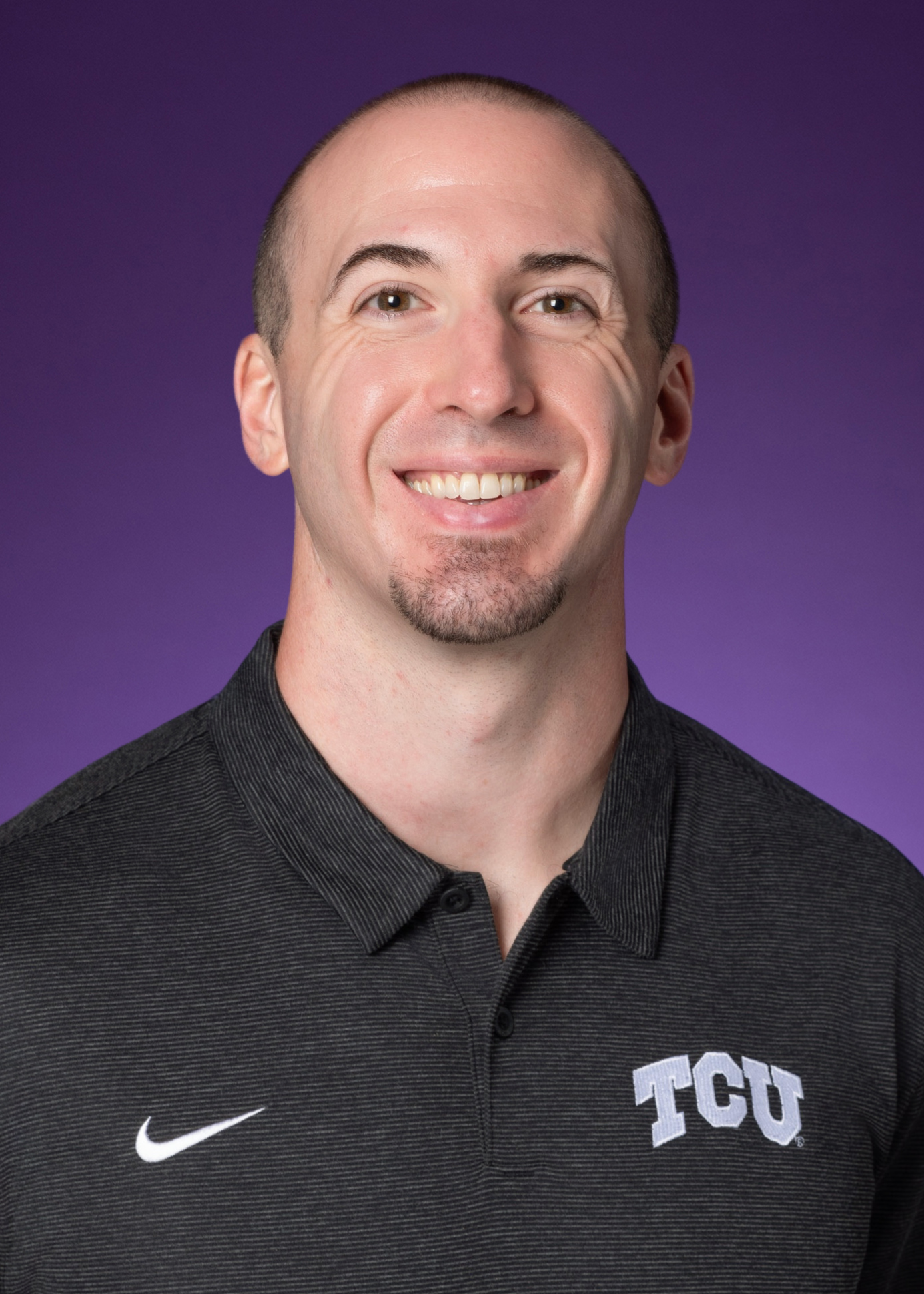 A photo of a man smiling, wearing a black polo shirt with a Nike logo on the left and the TCU logo on the right with a purple background.