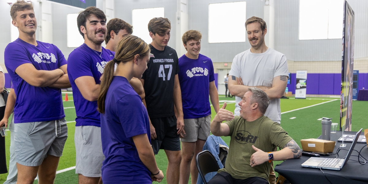 TCU Football players and a TCU Women's Soccer player stand around a booth at the Global Human Performance Forum inside the Sam Baugh Indoor Practice Facility