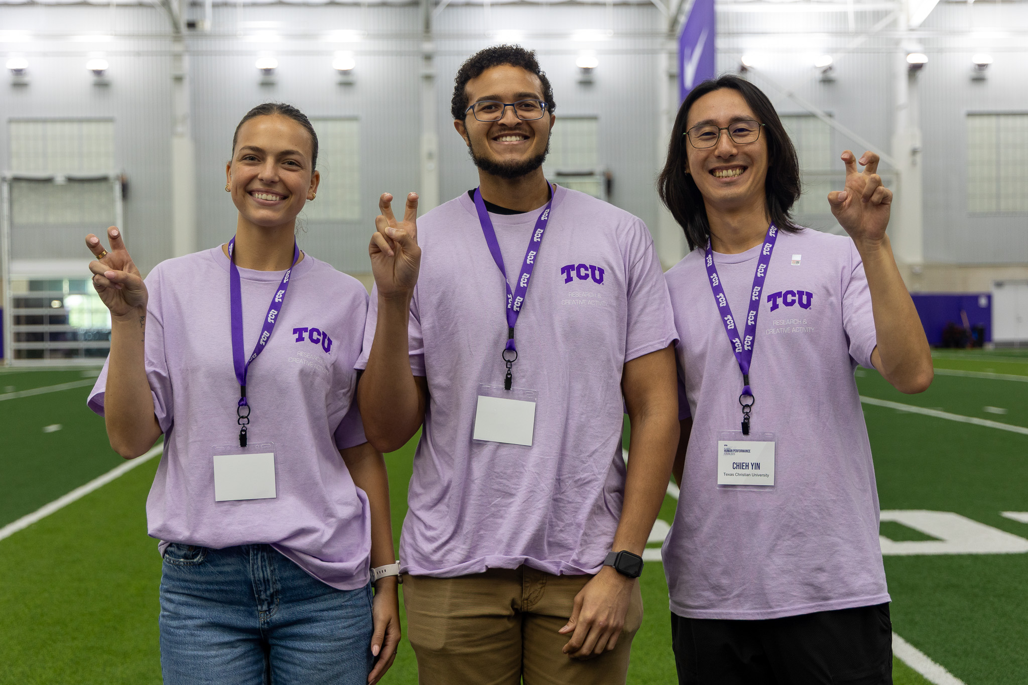 Three TCU Ph.D. students volunteer in the Sam Baugh Indoor Practice Facility at TCU during the first day of the Global Human Performance Forum. The three students are wearing matching purple shirts, purple lanyards, and throwing up the "Go Frogs" hand gesture.