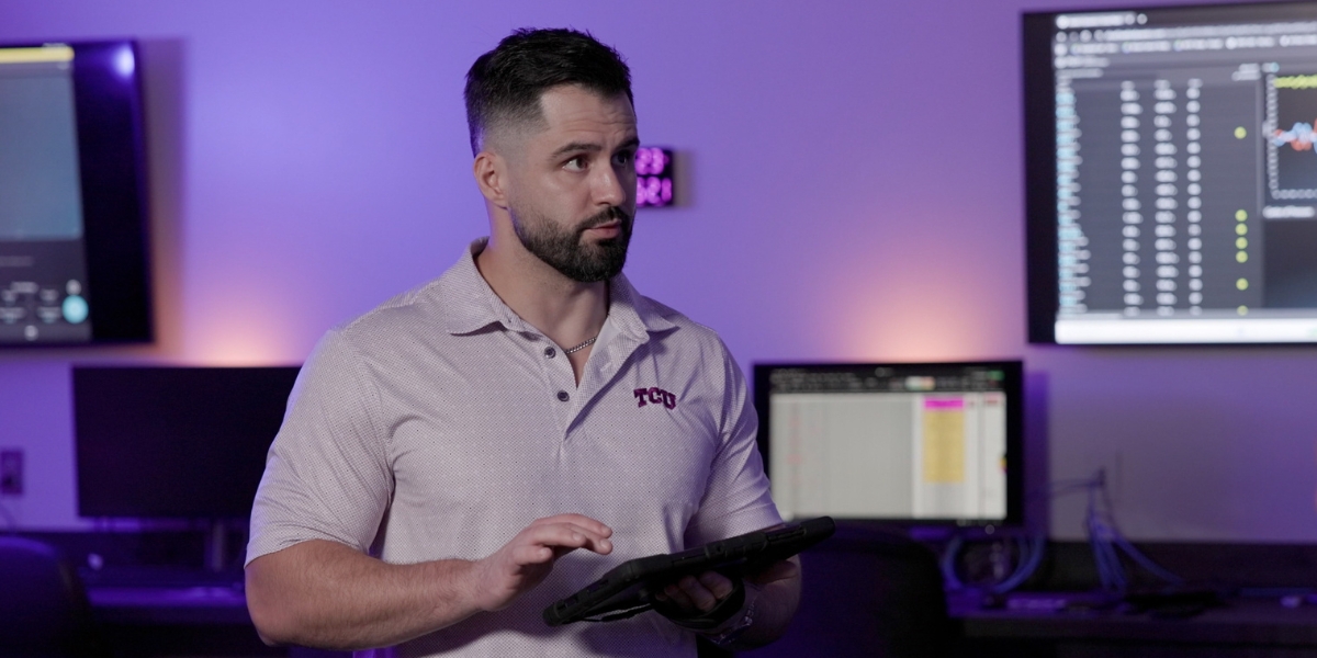 A photo of Austin Graybeal, wearing a TCU light purple polo shirt, holding an iPad. He is in the SHAPE Lab. Behind him are computer and TV monitors displaying data points and there is purple ambient lighting.