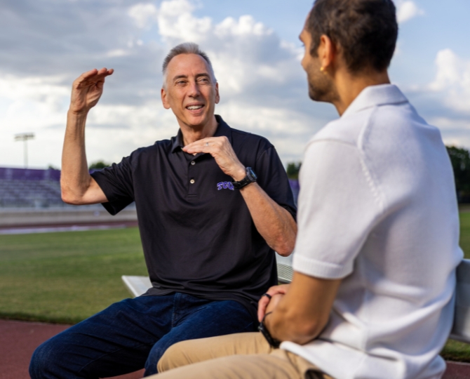 Two researchers sit on a bench beside TCU's outdoor track engaged in conversation. One gestures with both hands as if explaining a concept, while the other listens attentively. The running track, stadium seating and a cloudy evening sky provide the backdrop, emphasizing a discussion about athletics or biomechanics research.