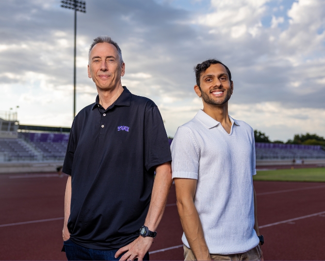 Two researchers stand side by side on an outdoor running track at TCU, smiling toward the camera. One wears a black TCU polo shirt, while the other wears a light gray short-sleeve knit polo. Stadium seating, track lanes and a dramatic cloudy sky form the background, highlighting the athletic research setting.