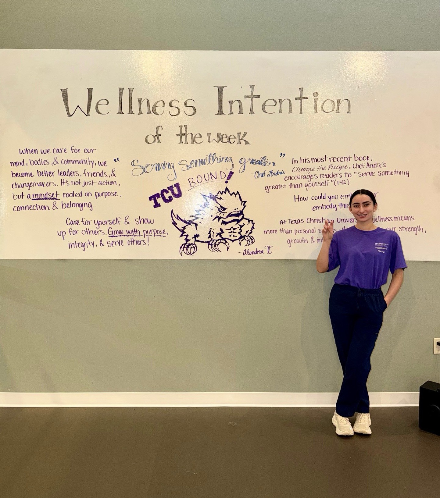 Alondra Trevizo stands in front of a white board that says "Wellness Intention of the week: Serving something greater" and the words "TCU Bound!" 