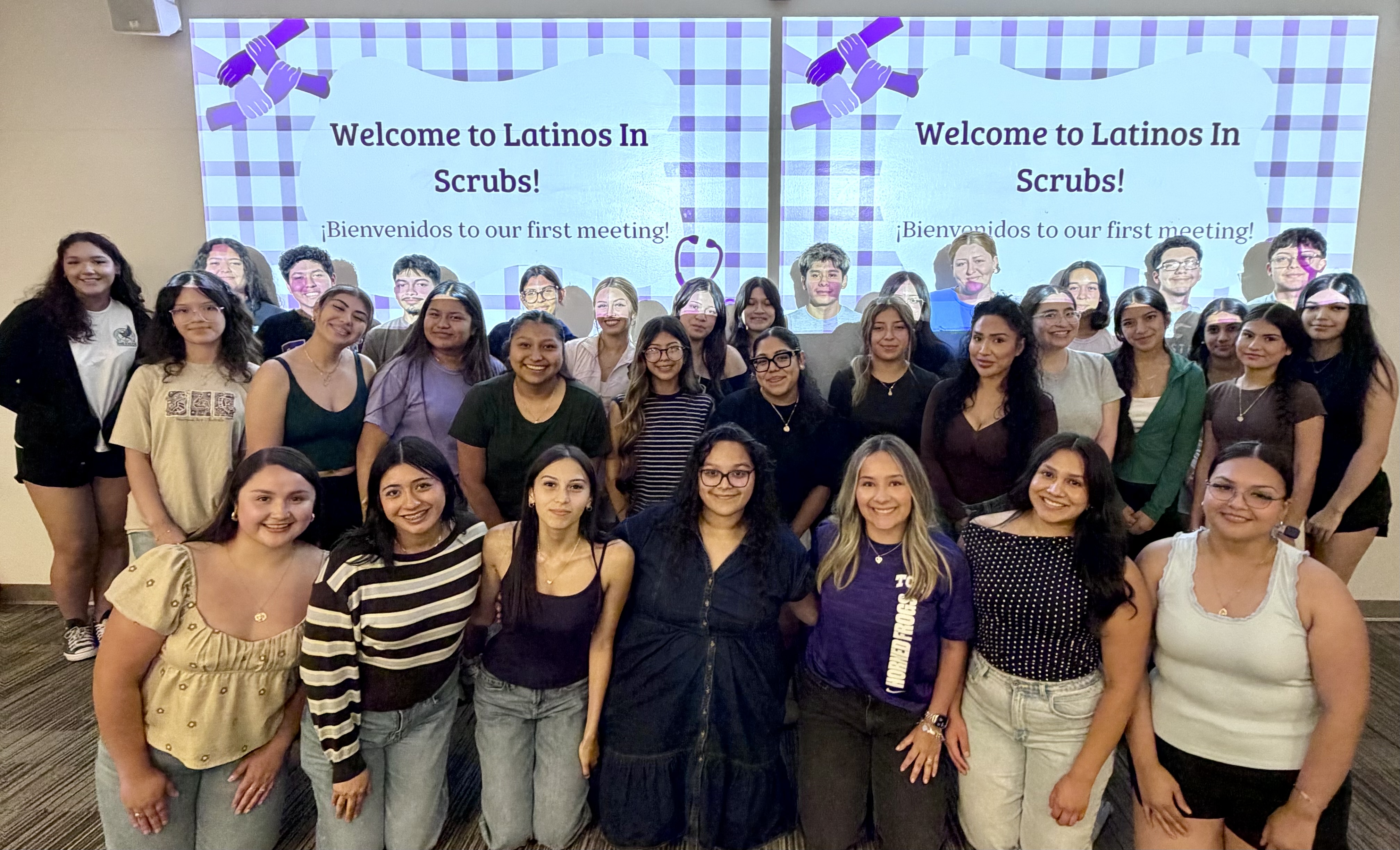 A group of more than two dozen women meet, standing and kneeling in front of a projection that says "Welcome to Latinos in Scrubs!" in English and Spanish in a classroom. 