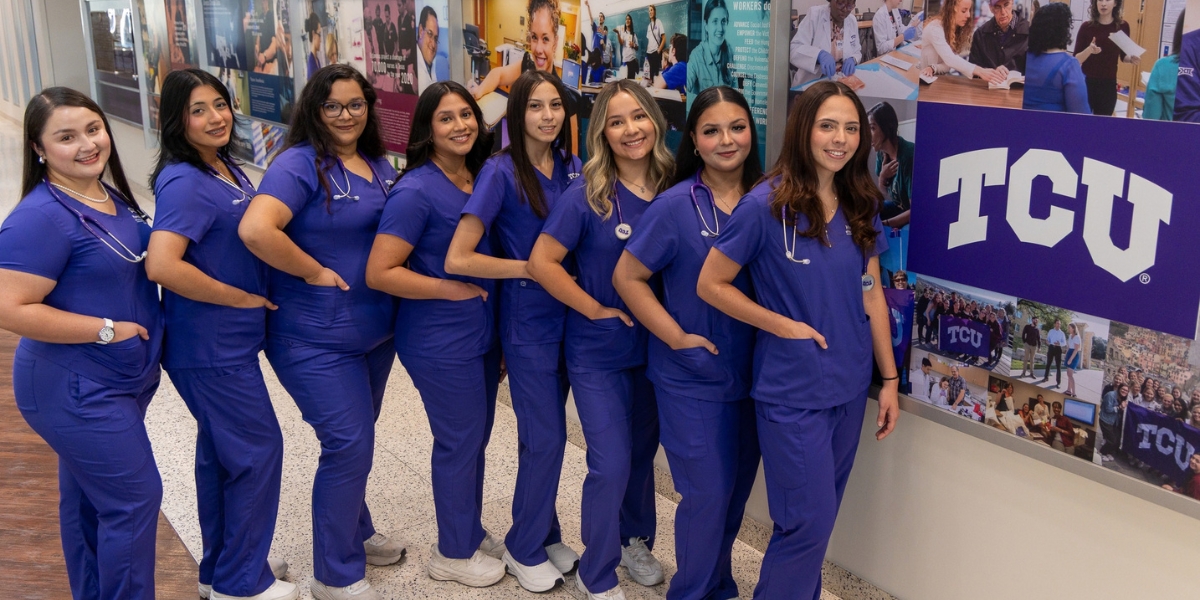 8 nursing students in purple scrubs stand in front of a wall with pictures and the arched TCU logo.