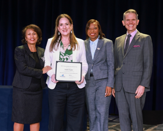 Four people pose together at an awards ceremony in front of a dark curtain. A woman wearing a white blazer over a green patterned blouse stands second from the left, smiling while holding a framed certificate. She is joined by two women in dark and gray business suits and a man in a gray suit with a purple shirt and tie. All four smile toward the camera in recognition of the award.