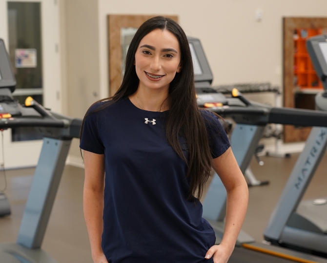 Close up image of Alondra Trevizo, standing in front of treadmills in a physical exercise space at a senior living community
