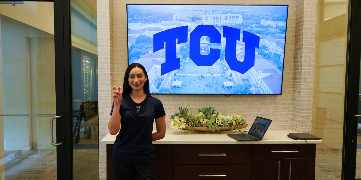 Alondra Trevizo standing in front of a large tv monitor with the abbreviation "TCU" on it. Her hand is in the "Go Frogs" gesture