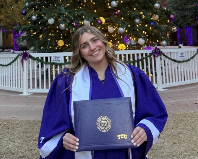 A woman smiling, wearing a TCU purple graduation gown and holding her TCU diploma cover. She stands in front of a Christmas tree on campus, surrounded by a small white fence.