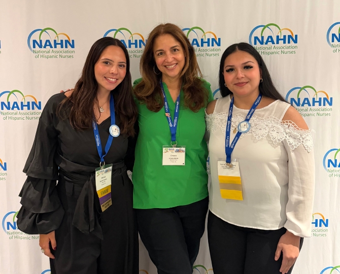 Dr. Griselle Batista, center, wears a green shirt and black pants with a blue lanyard. Leila Aguilar, right, is wearing a white long sleeve shirt with black pants and a blue lanyard. Monica Estefano, left, wears a black jumpsuit with a blue lanyard. The three women are standing in front of a white backdrop that says 