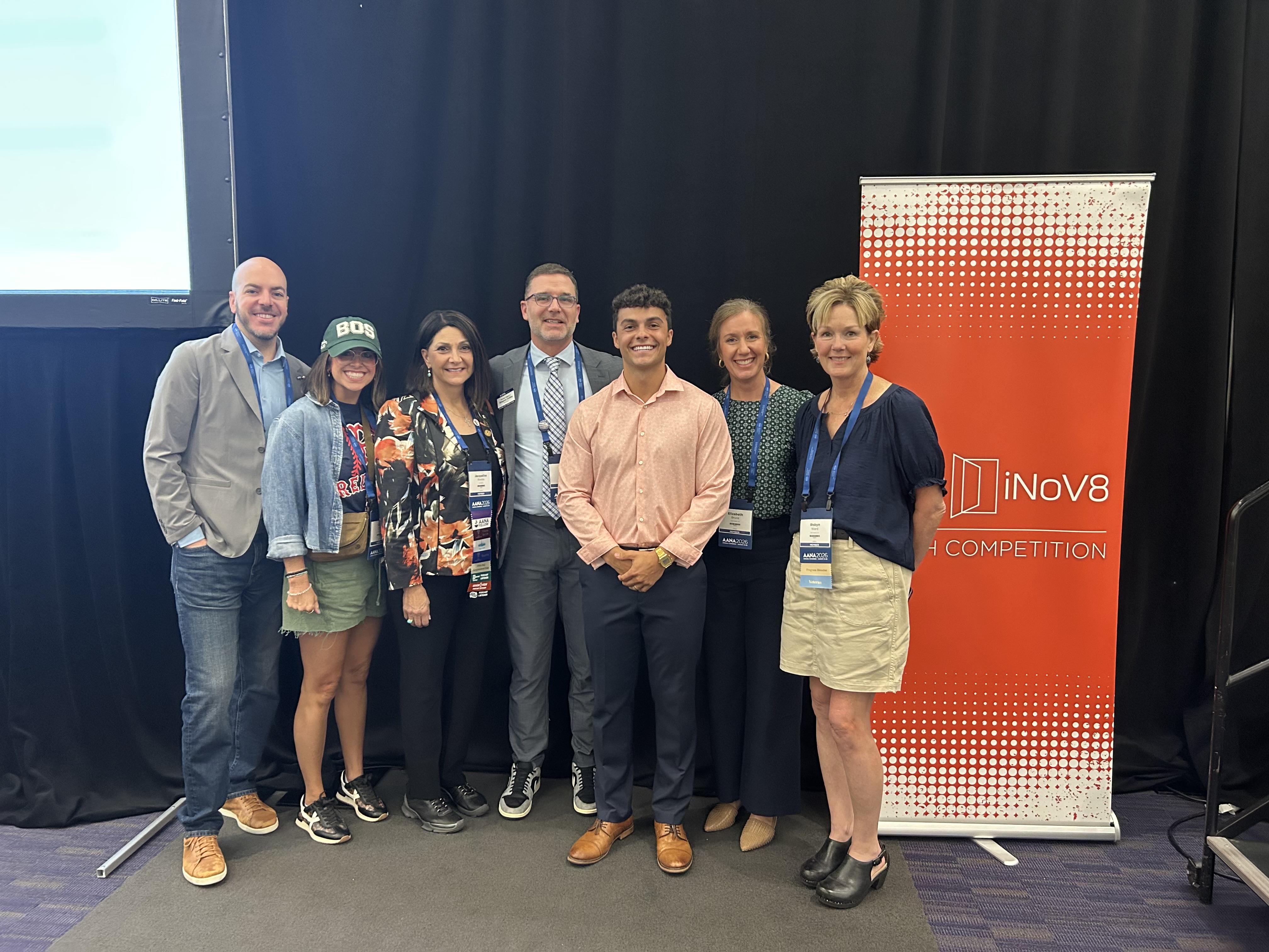Seven people stand together smiling for a group photo at a conference beside an orange and white "iNov8 Pitch Competition" banner. Several attendees wear conference badges, while one person is dressed casually in a baseball cap, denim jacket and shorts. The group poses in front of a black stage backdrop, suggesting they are participating in or attending the innovation competition.