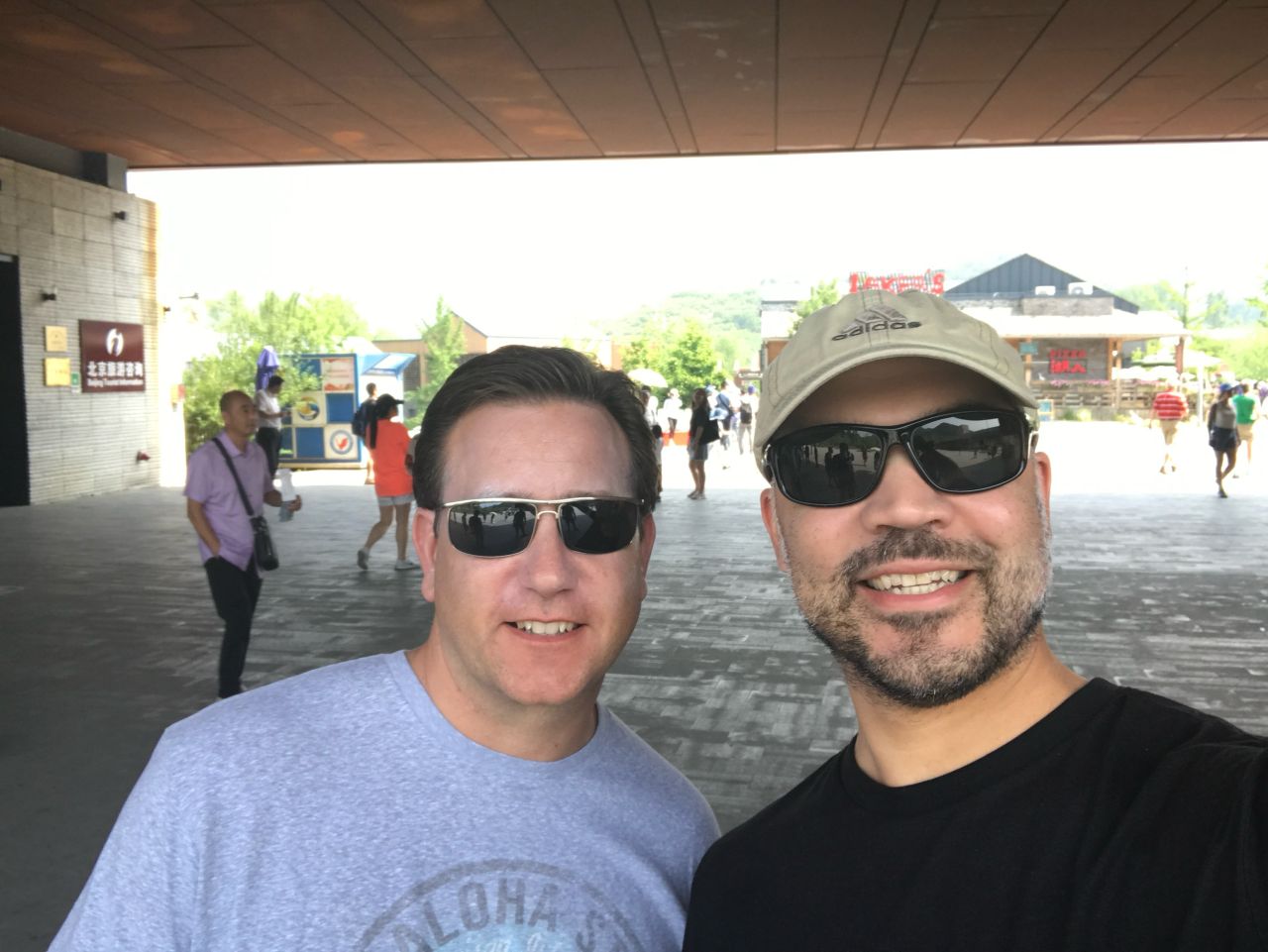 Two men wearing sunglasses smile for a selfie while visiting a public tourist area in China, with other visitors and buildings visible in the background.