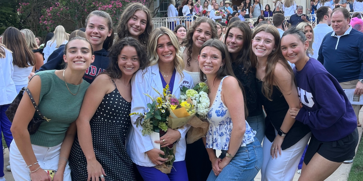 A nursing student in a white coat holds bouquets of flowers while posing with a large group of smiling friends outdoors during a White Coat Ceremony celebration on the TCU campus.
