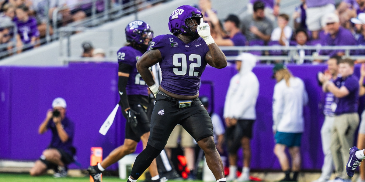 TCU defensive tackle Ansel Din-Mbuh, wearing a purple No. 92 football jersey, black pants and a purple helmet, stands on the field with one hand on his hip and his other hand raised to his facemask in a celebratory gesture. A teammate, photographers and cheering fans are visible in the background at TCU's football stadium.