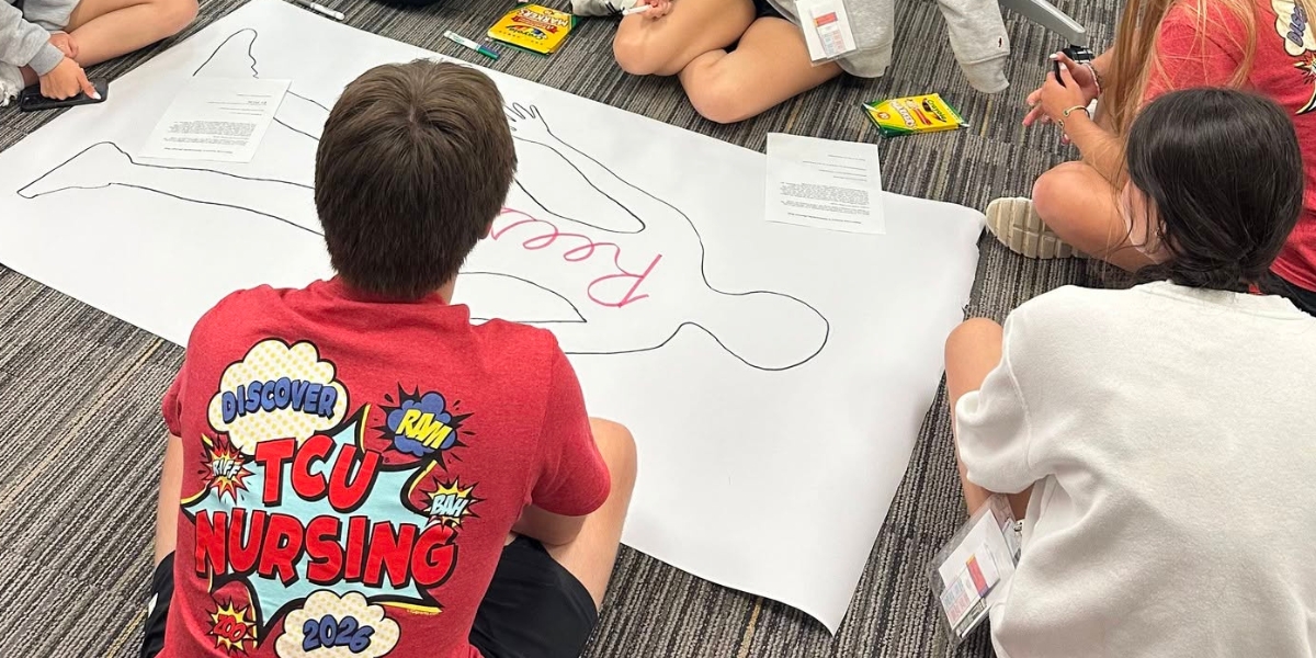 Several high school students are sitting on the ground, circled around a life-size outline of a person as a "patient" for a coloring activity. The students are wearing red shirts that say "TCU Nursing 2026"